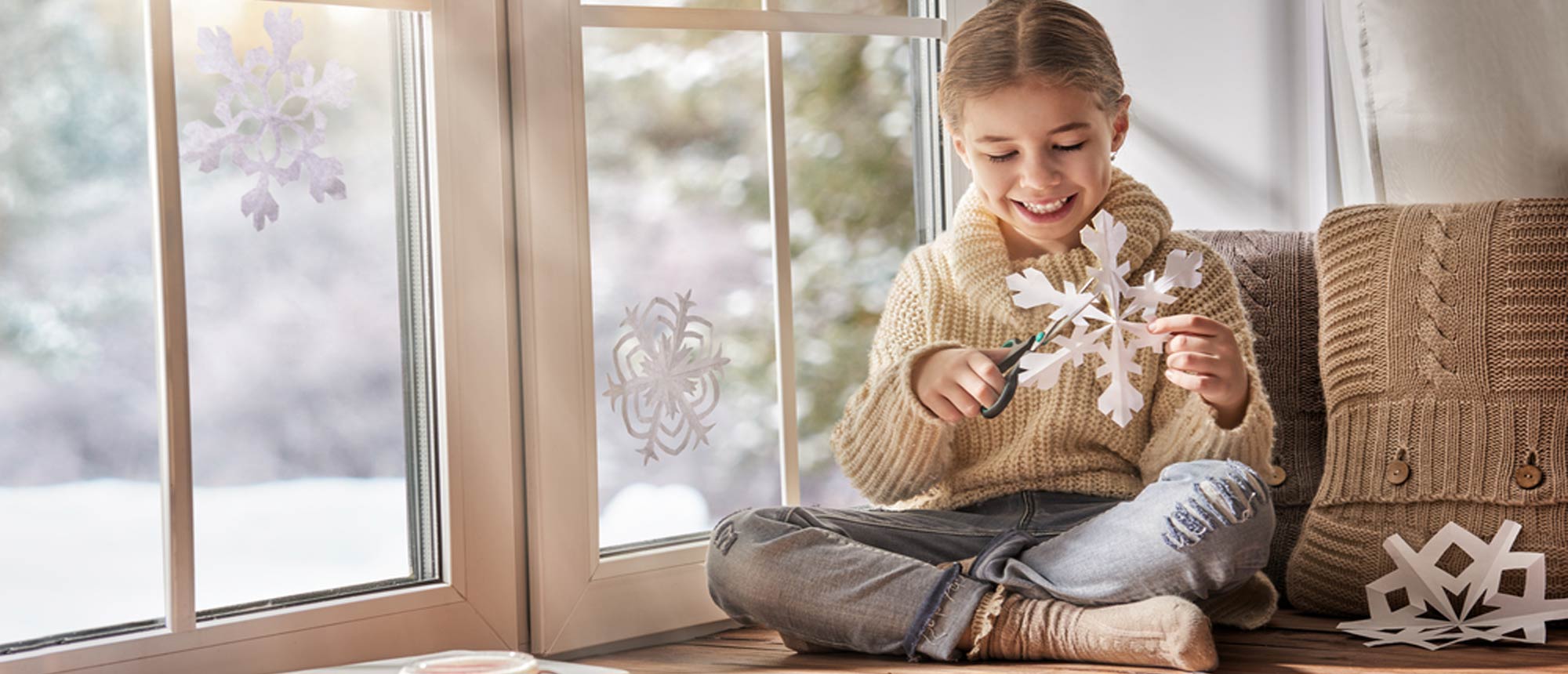 Young girl making paper snow flakes while sitting by a window showing snow outside